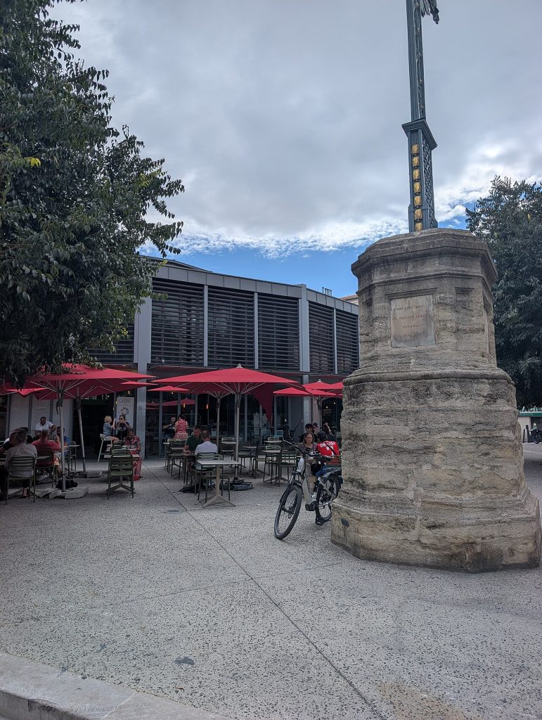 Des terrasses aux parasols rouges devant la halle Laissac, près de la croix de Villaret