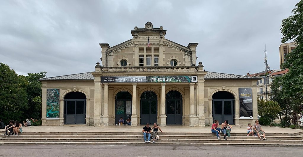 Le Pavillon Populaire, pavillon à colonnes au fronton sculpté, avec des passants assis sur ses marches