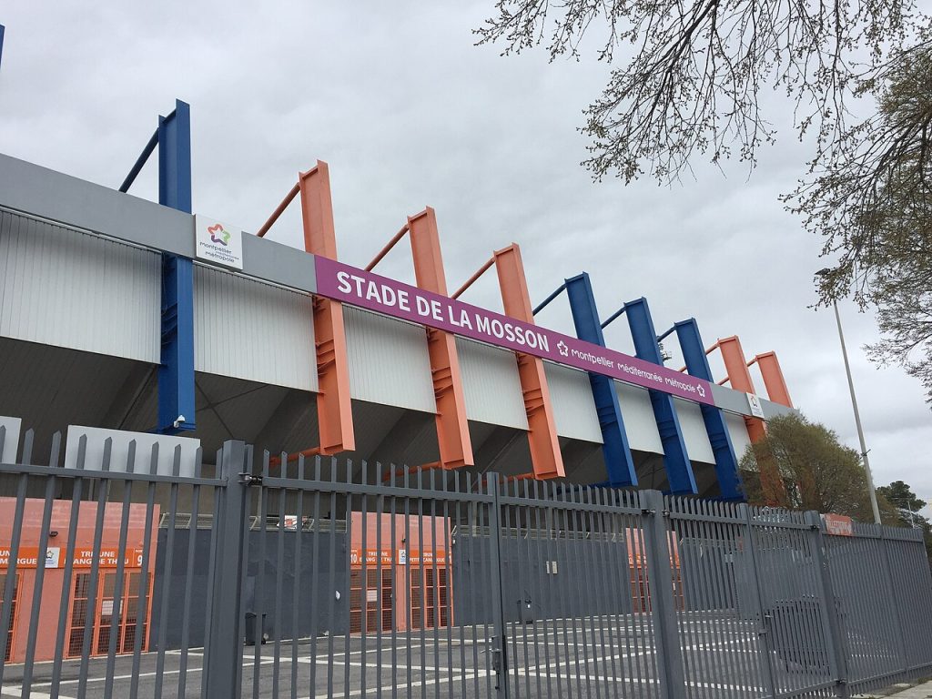 La tribune du stade de la Mosson, charpente de poutres métalliques orange, bleues et rouges, et son bandeau « Stade de la Mosson »