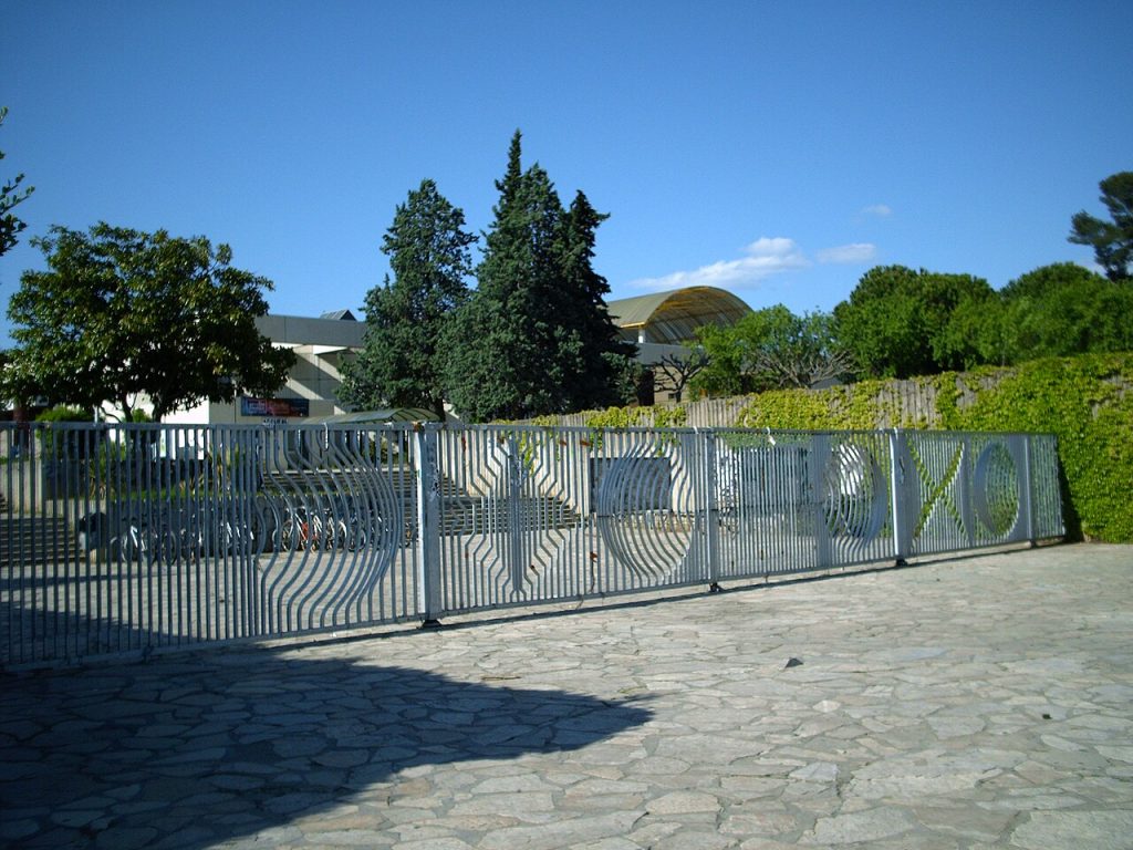 Le portail d’entrée de l’université Paul-Valéry, grille métallique ajourée, sous les pins et un ciel bleu