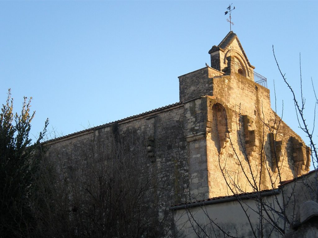 L’église Sainte-Croix de Celleneuve : haut mur de pierre doré par le soleil, arcs défensifs et clocher-arcade surmonté d’une girouette