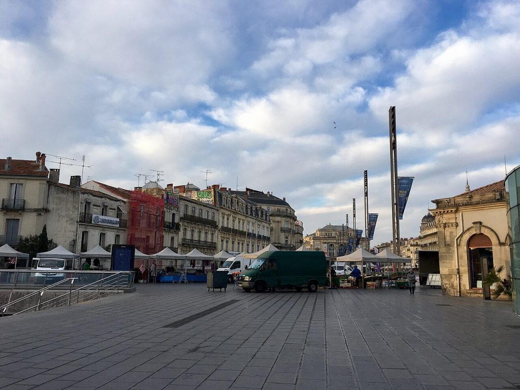Stands blancs du marché installés sur la place de la Comédie, un matin nuageux, devant une rangée d’immeubles anciens