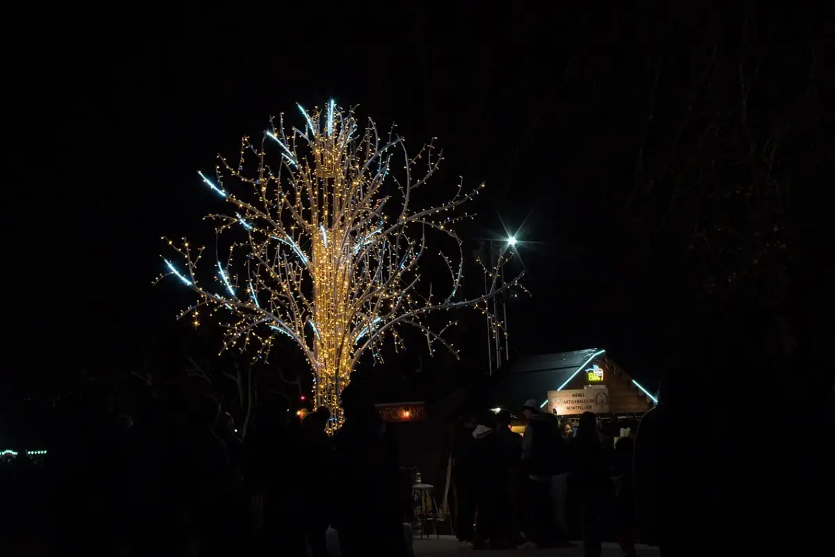 Un arbre lumineux et un chalet éclairé au marché de Noël de Montpellier, de nuit, avec des visiteurs en ombre chinoise