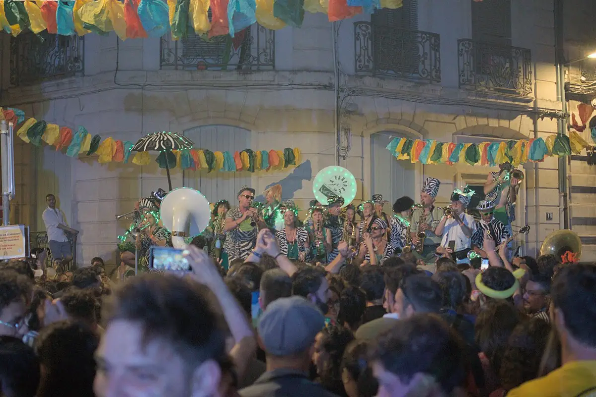 Une fanfare en costumes joue le soir sous des guirlandes de papier, devant une foule dense, dans une rue du quartier Beaux-Arts